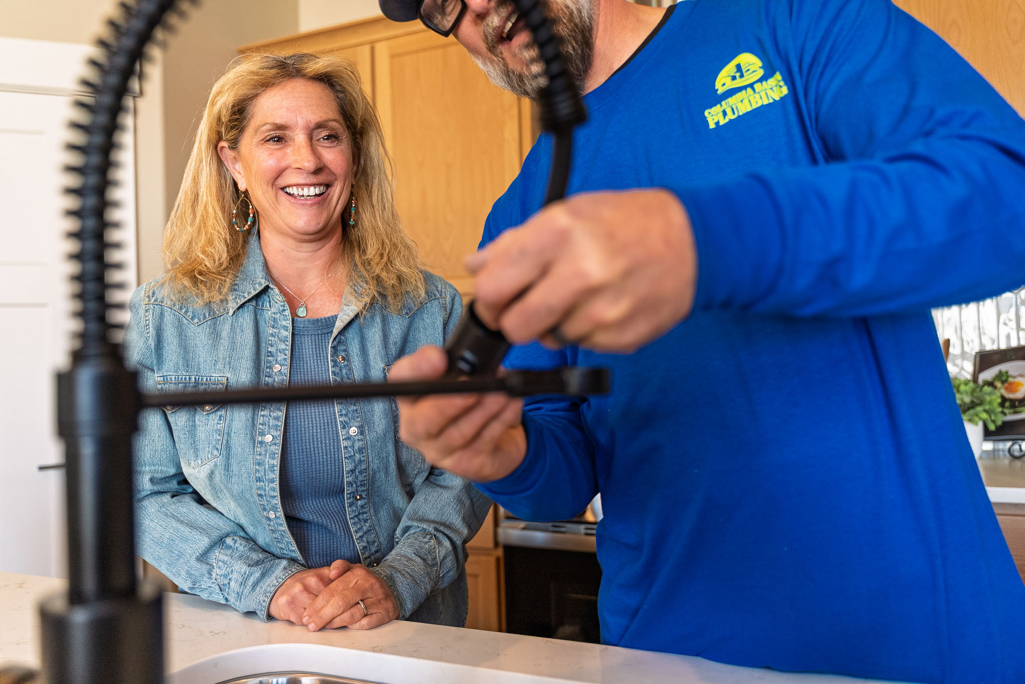 Plumber fixing kitchen faucet with woman watching, smiling.